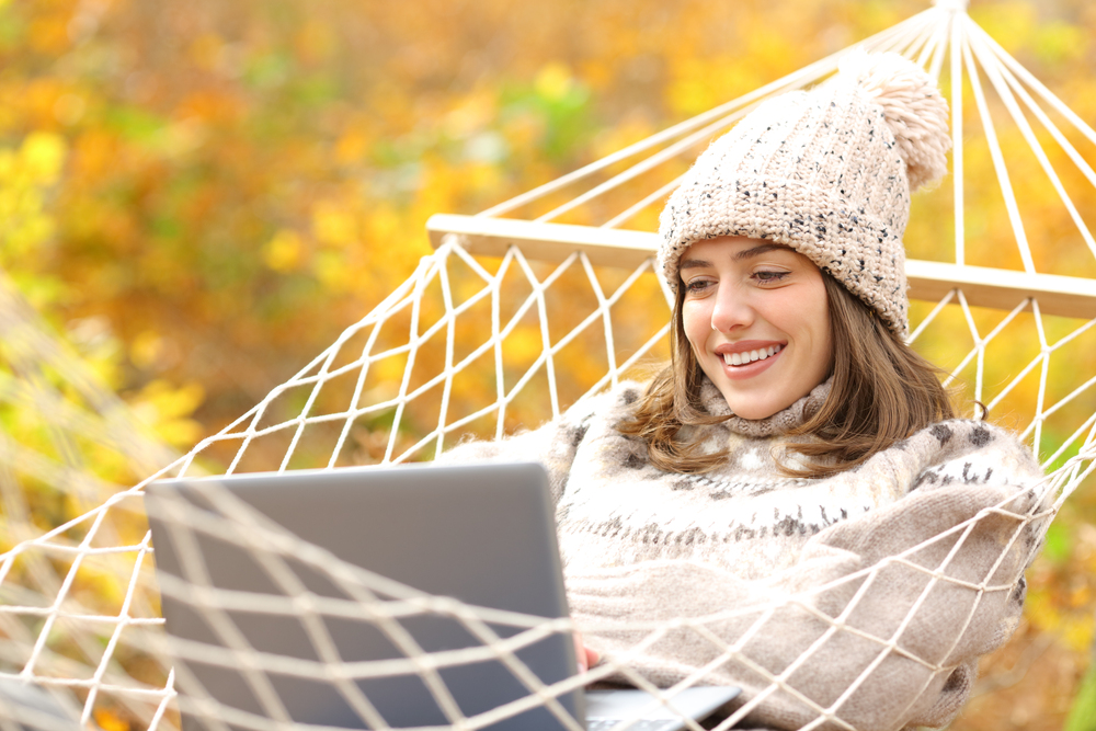 Glückliche Frau liegt in einer Hängematte und benutzt einen Laptop im Herbst in einem Wald.