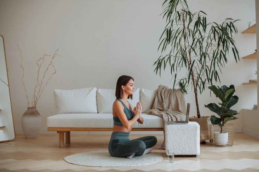 Eine junge brünette Frau in einem Trainingsanzug macht Yoga in einer Online-Lektion auf einem Laptop zu Hause im Wohnzimmer. Sie sitzt im Lotussitz mit geschlossenen Augen. Im Raum steht ein weißes Sofa.